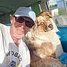 man, dog, selfie, white_cap, sunglasses, golden_brown_dog, fluffy_dog, vehicle_interior, blue_seat, green_curtain, white_tshirt, sunlight, portrait, closeup, companion, pet, casual, smile, beard, middle_aged