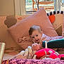 baby, child, pink_pillow, toy, indoor, window, cushion, smiling, face, floor, play_area, soft_toys, furniture, natural_light, wood_floor, clothing, person, happy, home, relaxing