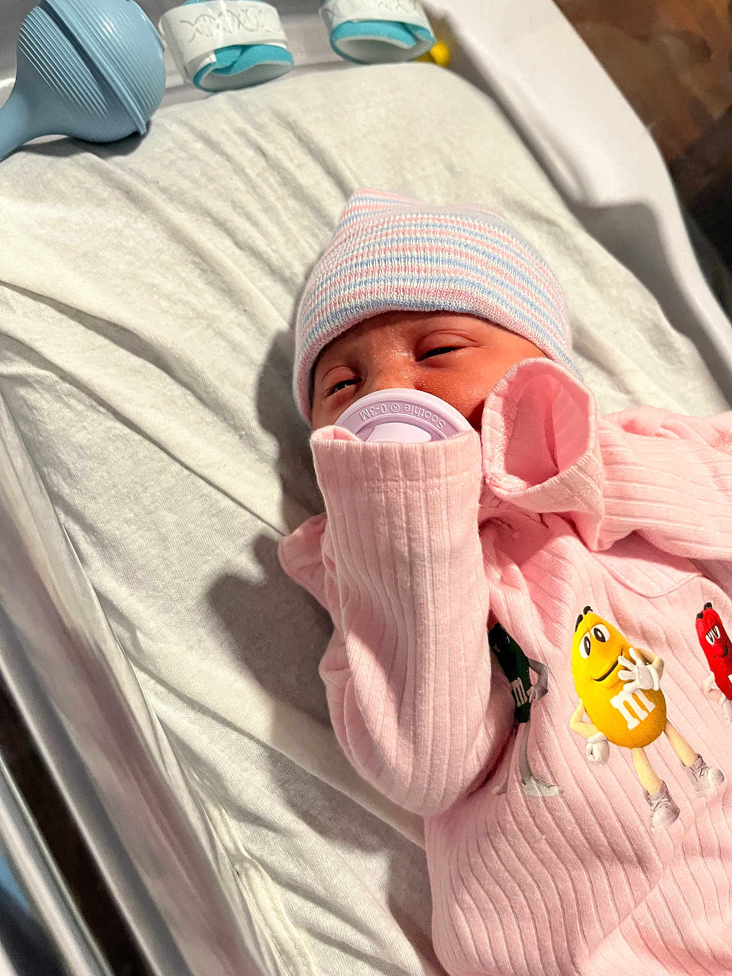 Marissa is registered to the contest to win money with this photo: newborn, baby, pacifier, pink_clothing, hat, striped_hat, m_and_ms, onesie, bed, blanket, crib, infant, sleepy, closeup, person, soft_lighting, indoors, cute, baby_toys, sleep