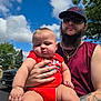 Charles is registered to the contest to win money with this photo: baby, man, outdoor, sky, clouds, trees, tattoo, beard, sunglasses, red_clothing, holding, person, child, parking_lot, vehicle, daylight, nature, casual, portrait, summer
