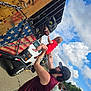 Charles is registered to the contest to win money with this photo: baby, man, tattoos, cap, red_clothing, wooden_crate, american_flag, dinosaur_head, football, metal_bars, outdoor, cloudy_sky, people, crowd, parking_lot, vehicle, chain, playful, fun, interaction