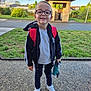 child, glasses, backpack, toy, smile, outdoor, sidewalk, grass, building, jacket, sneakers, person, young, happy, casual_clothing, daylight, nature, headwear, portrait, footwear