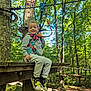 child, smiling, outdoor, forest, trees, wood, platform, harness, rope_course, adventure, sneakers, greenery, sunlight, nature, playground, young_child, casual_clothing, safety_gear, daylight, happy