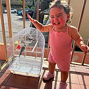 Izabelle is registered to the contest to win money with this photo: child, toddler, pink_clothing, birdcage, bird, balcony, sunlight, smile, happy, curly_hair, shoes, outdoor, railing, playful, person, daylight, expression, young_child, fence, building