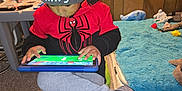 Vincent is registered to the contest to win money with this photo: beanie, bed, carpet, casual, child, focus, furniture, gray_pants, indoor, kid, person, playing, red_hoodie, seat, sneakers, spiderman, tablet, technology, toys, wood_paneling