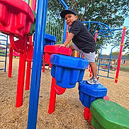 Vincent is registered to the contest to win money with this photo: child, boy, playground, climbing, cap, black_shirt, shorts, sneakers, play_equipment, colorful, outdoor, daylight, tree, wood_chips, park, fun, active, smiling, recreation, summer