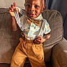 toddler, child, boy, suspenders, bow_tie, shirt, pants, smile, face, hand, couch, indoors, curly_hair, window, sunlight, fabric, home, portrait, playful, casual_clothing