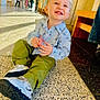 child, toddler, smiling, indoor, hallway, floor, toy, blond_hair, casual_clothing, green_pants, blue_shirt, sneakers, people_in_background, natural_light, benches, tile_floor, happy, sitting, portrait, daylight