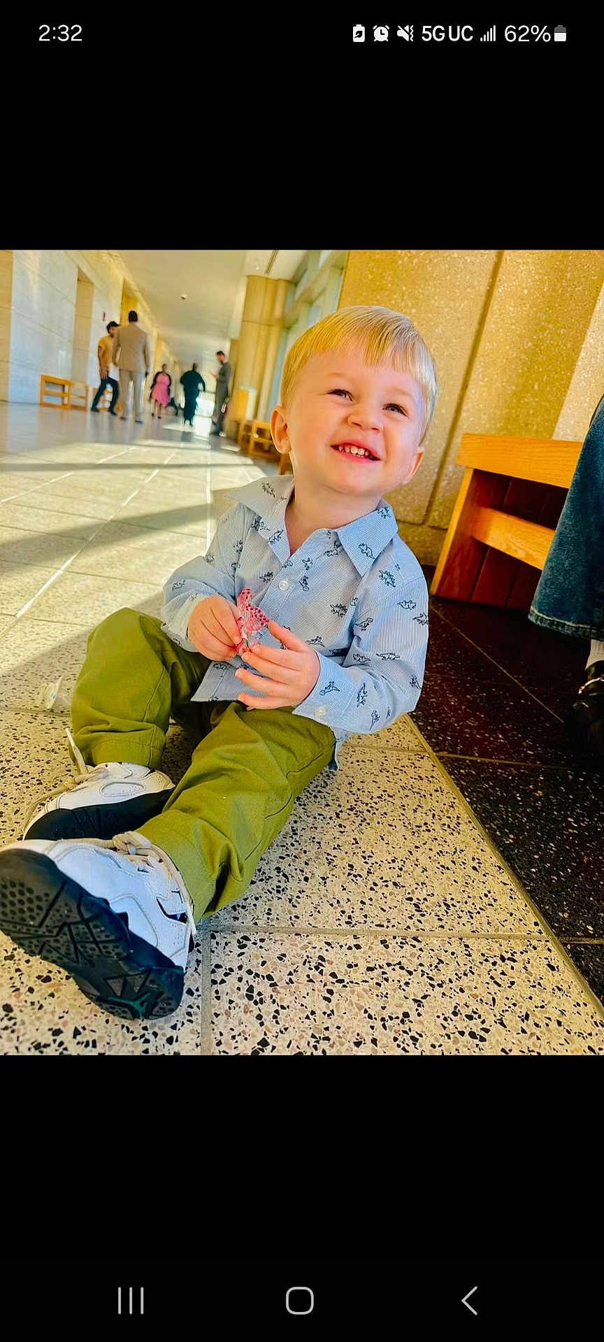 Brian is registered to the contest to win money with this photo: child, toddler, smiling, indoor, hallway, floor, toy, blond_hair, casual_clothing, green_pants, blue_shirt, sneakers, people_in_background, natural_light, benches, tile_floor, happy, sitting, portrait, daylight