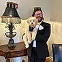 Paddington joined the competition — help win amazing prizes! dog, puppy, person, tuxedo, bow_tie, glasses, beard, decorative_lamp, wooden_table, furniture, indoor, holding, pet, smiling, lampshade, armchair, beige_wall, formal_wear, floor_tiles, portrait