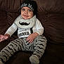Elijah joined the competition — help win amazing prizes! baby, child, smile, hat, bib, pants, mustache_pattern, sitting, couch, brown, clothing, person, indoor, cute, infant, young_child, text_on_clothing, reaching_hand, portrait, cozy