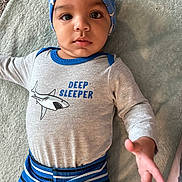 Elijah joined the competition — help win amazing prizes! baby, child, infant, blue_cap, striped_pants, grey_shirt, deep_sleeper_text, cartoon_shark, lying_down, soft_surface, curious_expression, large_eyes, hand_reaching, indoor, close_up, cute, portrait, clothing, headwear, person