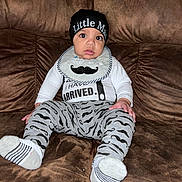 Elijah is registered to the contest to win money with this photo: baby, child, infant, couch, brown_couch, clothing, hat, bib, mustache_pattern, pants, socks, sitting, portrait, indoor, cute, wide_eyes, looking_at_camera, person, young_child, casual