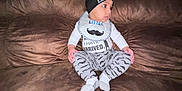 Elijah is registered to the contest to win money with this photo: baby, infant, child, couch, brown_couch, clothing, hat, bib, mustache_pattern, socks, sitting, indoors, looking_sideways, cute, cozy, comfortable, person, young_child, casual_wear, portrait