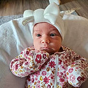 Cassi is registered to the contest to win money with this photo: baby, infant, face, hat, white_hat, onesie, floral_pattern, blanket, indoors, portrait, cute, child, young, soft_texture, cozy, sleepwear, expression, person, newborn, skin