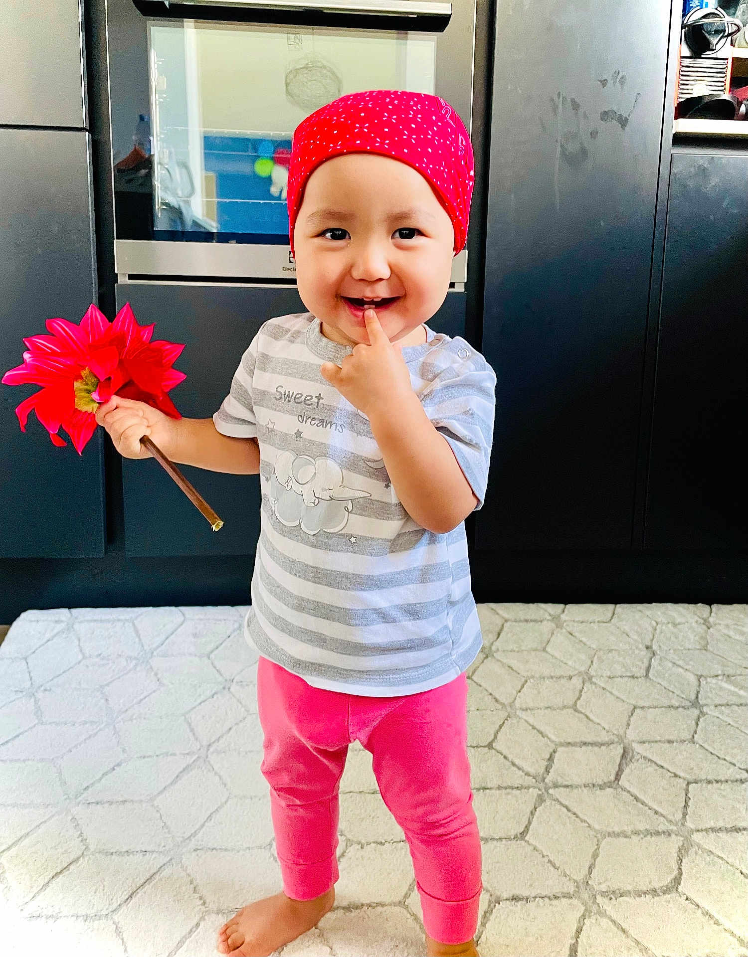 Maya a rejoint le concours — aidez-le/la à gagner de superbes lots ! toddler, child, red_bandana, flower, pink_pants, striped_shirt, barefoot, indoor, kitchen, rug, oven, cabinet, smiling, playful, person, standing, hand, finger, cute, home