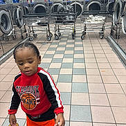 Nolan is registered to the contest to win money with this photo: child, laundromat, washing_machine, laundry_cart, tiled_floor, red_sweatshirt, orange_shorts, sneaker, curious, indoor, floor_tiles, reflection, basket, laundry, clothing, young_child, person, portrait, casual, head