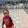 child, laundromat, washing_machine, laundry_cart, tiled_floor, red_sweatshirt, orange_shorts, sneaker, curious, indoor, floor_tiles, reflection, basket, laundry, clothing, young_child, person, portrait, casual, head