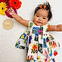baby, child, infant, colorful_dress, flowers, gerbera_daisies, wooden_plaque, four_months, smiling, cute, lying_down, hair_clips, white_background, patterned_clothing, happy, portrait, young_child, outstretched_arms, indoor, playful