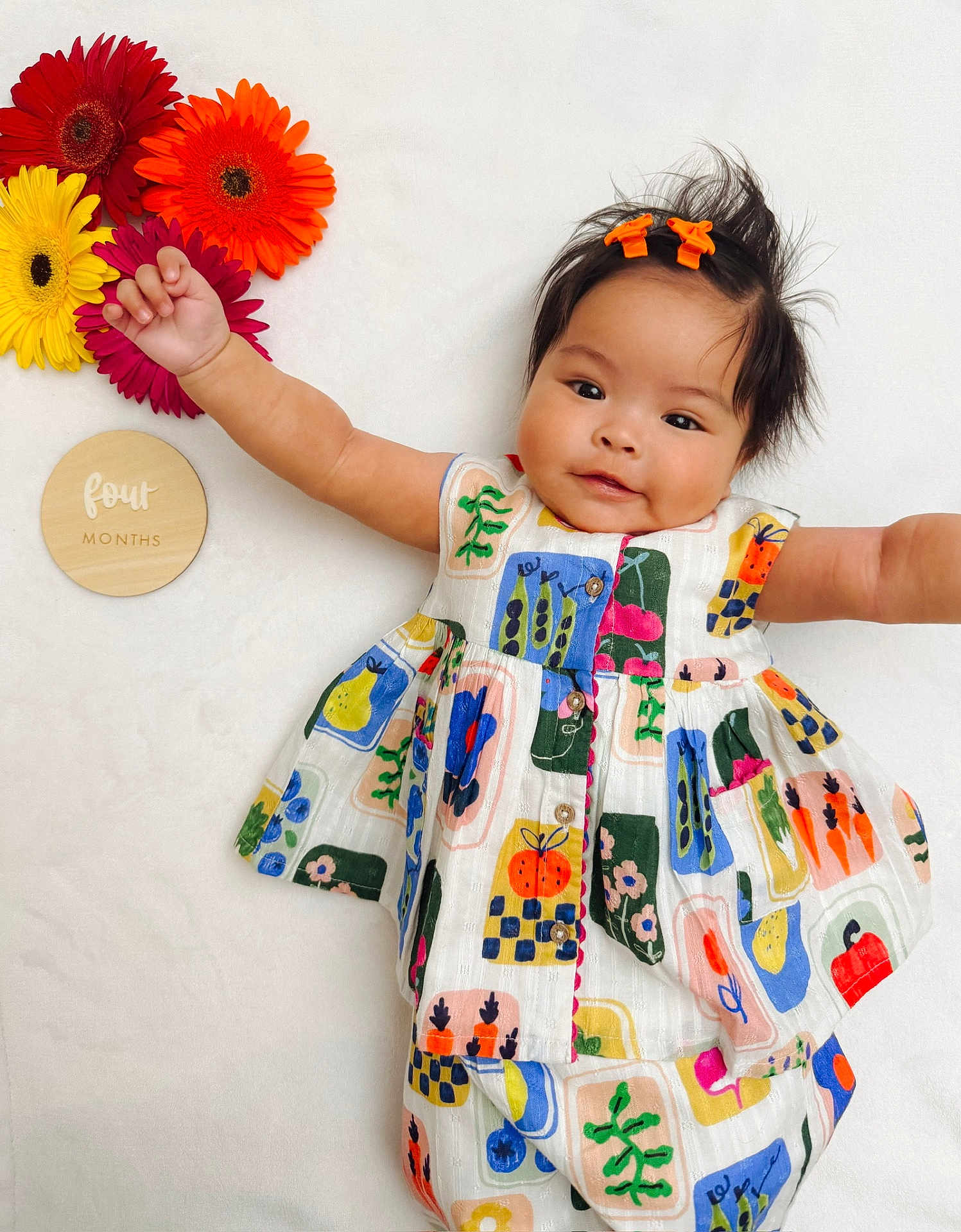 Gianna is registered to the contest to win money with this photo: baby, child, infant, colorful_dress, flowers, gerbera_daisies, wooden_plaque, four_months, smiling, cute, lying_down, hair_clips, white_background, patterned_clothing, happy, portrait, young_child, outstretched_arms, indoor, playful
