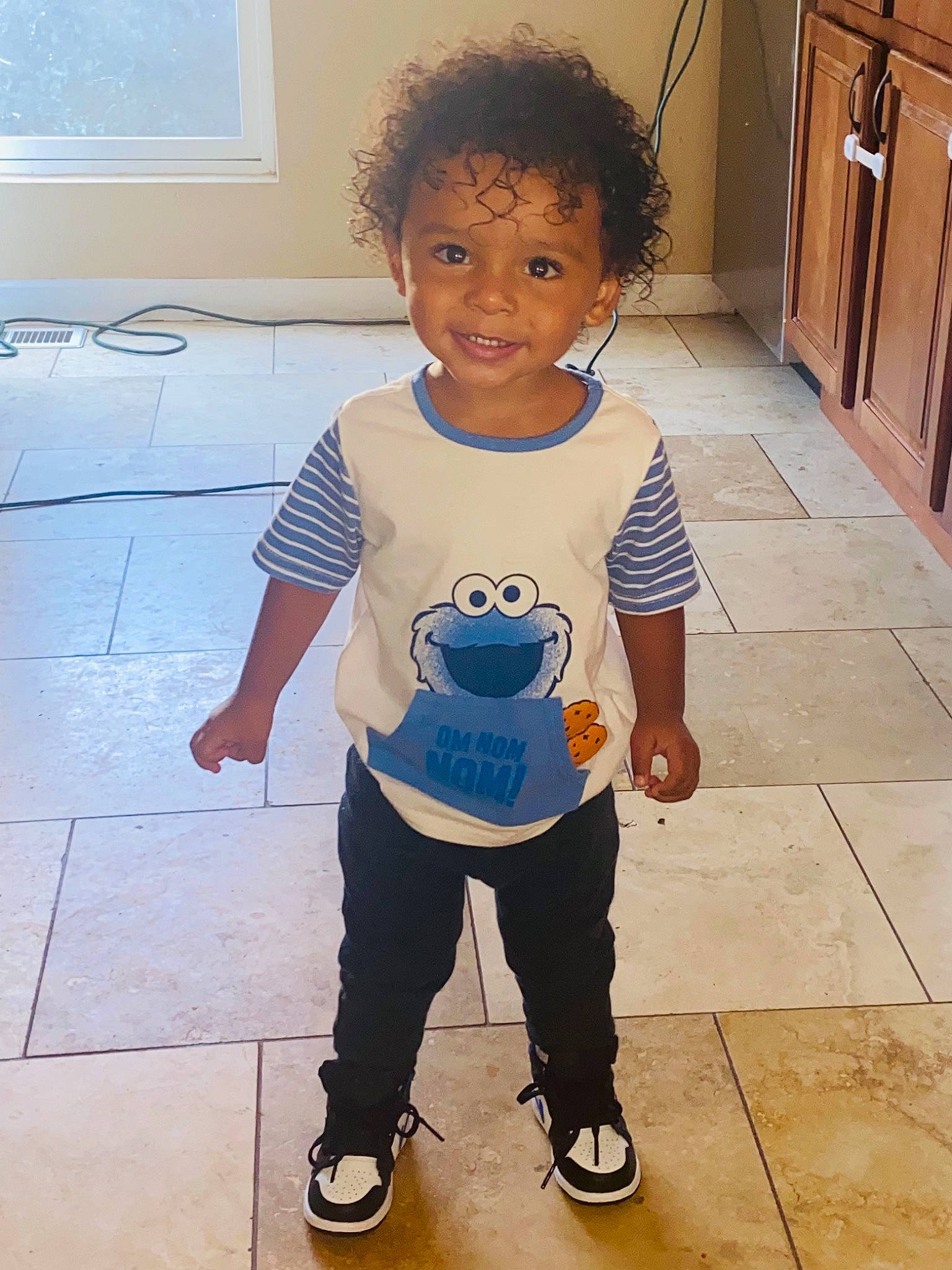 Harlem is registered to the contest to win money with this photo: baby_toddler_clothing, cabinetry, child, cupboard, eye, floor, flooring, hair, hairstyle, happy, head, human_body, joint, joy, leg, person, sleeve, smile, standing, t_shirt