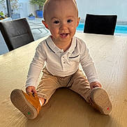 Hugo participe au concours pour gagner de l'argent avec cette photo : baby, child, smiling, happy, table, wooden_table, indoor, window, pool, pants, shirt, footwear, brown_shoes, person, face, seated, home, daylight, plant, background