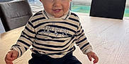 Hugo participe au concours pour gagner de l'argent avec cette photo : baby, child, indoor, table, wooden_table, striped_clothing, brown_shoes, smiling, sitting, curious, face, person, window, daylight, furniture, home, pants, shoe, cute, happy