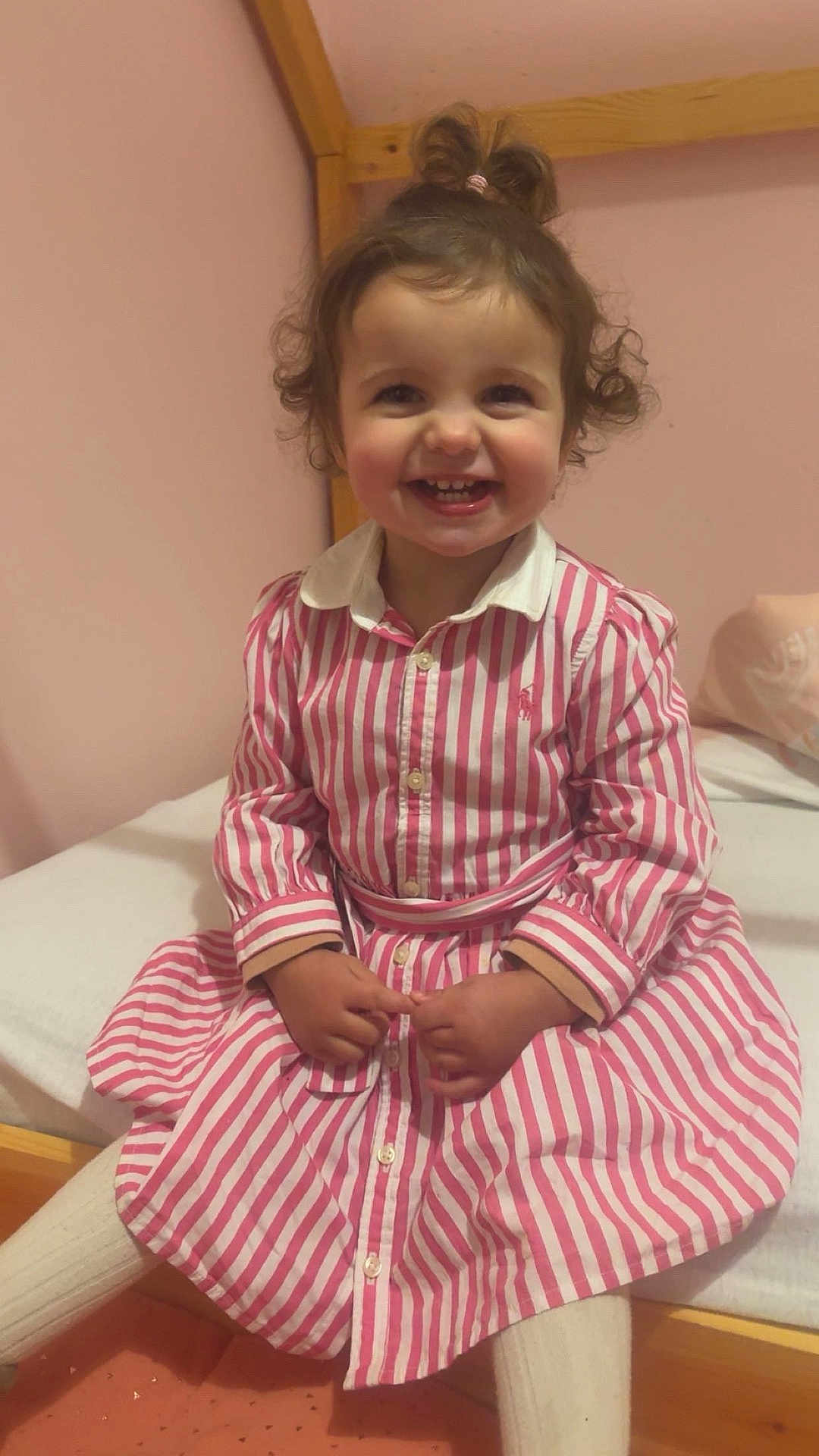 Ambre a rejoint le concours — aidez-le/la à gagner de superbes lots ! toddler, child, smiling, curly_hair, pink_dress, striped_clothing, white_tights, indoor, bed, wooden_frame, happy, person, portrait, cute, sitting, young_child, playful, room, cozy, baby