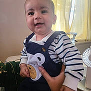 Qasim is registered to the contest to win money with this photo: baby, child, smiling, striped_shirt, overalls, bear_patch, hand, indoor, curtains, fan, person, happy, cute, infant, portrait, clothing, face, holding, bright, window