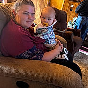 Kace is registered to the contest to win money with this photo: woman, baby, pacifier, recliner, indoor, wood_paneling, plaid_shirt, braided_hair, casual_clothing, living_room, man, jeans, black_shirt, portrait, family, comfort, home, furniture, carpet, table
