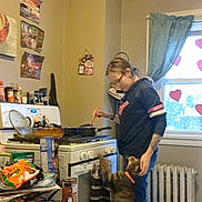 Krueger is registered to the contest to win money with this photo: person, cat, kitchen, stove, cooking, window, curtains, snacks, table, radiator, floor, glasses, sweatshirt, cat_collar, pots, pans, wall_art, oven_mitts, heart_decorations, indoors
