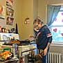 person, cat, kitchen, stove, cooking, window, curtains, snacks, table, radiator, floor, glasses, sweatshirt, cat_collar, pots, pans, wall_art, oven_mitts, heart_decorations, indoors