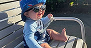 Luka joined the competition — help win amazing prizes! baby, child, sunglasses, hat, bucket_hat, smile, outdoor, bench, sunlight, pacifier, blue_clothing, shark_pattern, happy, summer, shade, feet, hand, metal_bench, playful, cute