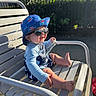 baby, child, sunglasses, hat, bucket_hat, smile, outdoor, bench, sunlight, pacifier, blue_clothing, shark_pattern, happy, summer, shade, feet, hand, metal_bench, playful, cute