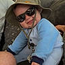 Luka is registered to the contest to win money with this photo: baby, child, sunglasses, hat, blue_onesie, smiling, indoor, couch, remote_control, person, cozy, cute, infant, funny, hat_strap, relaxed, home, adult_leg, playful, casual
