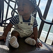 Zhakari is registered to the contest to win money with this photo: baby, child, smiling, hat, striped_clothing, overalls, socks, chair, balcony, ocean, sky, sunlight, happy, portrait, outdoor, nautical, infant, seat, person, daytime