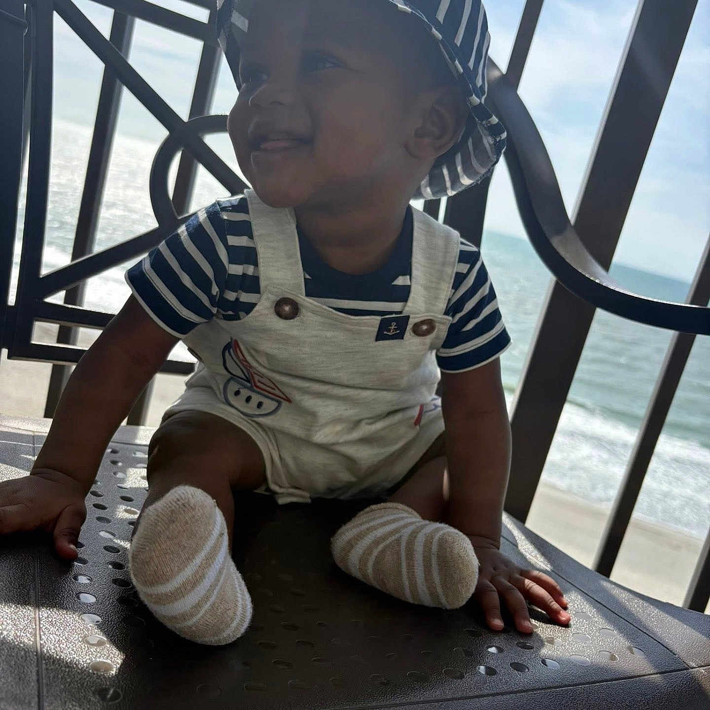 Zhakari is registered to the contest to win money with this photo: baby, child, smiling, hat, striped_clothing, overalls, socks, chair, balcony, ocean, sky, sunlight, happy, portrait, outdoor, nautical, infant, seat, person, daytime