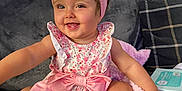 Lottie is registered to the contest to win money with this photo: baby, child, smiling, sitting, couch, pink_bow, headband, floral_dress, white_socks, cushion, remote_control, baby_wipes, indoor, happy, cute, portrait, infant, home, comfort, playful