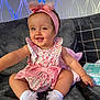 baby, child, smiling, sitting, couch, pink_bow, headband, floral_dress, white_socks, cushion, remote_control, baby_wipes, indoor, happy, cute, portrait, infant, home, comfort, playful