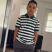 Yadiel Núñez is registered to the contest to win money with this photo: boy, child, indoor, striped_shirt, polo_shirt, pants, standing, neutral_expression, door, window_blinds, armchair, home, floor, person, casual_clothing, portrait, young, furniture, living_room, hands_in_pockets