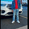 child, boy, smiling, denim_jacket, red_shirt, jeans, sneakers, car, white_car, parking_lot, outdoor, daytime, asphalt, buildings, residential, trees, casual_clothing, happy, person, portrait