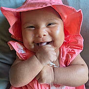 Izzy is registered to the contest to win money with this photo: baby, infant, child, smiling, happy, cute, pink_hat, pink_dress, hand, face, skin, indoors, portrait, closeup, young_child, cheeks, sitting, cushion, bright_colors, joy