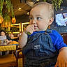 toddler, child, baby, overalls, blue_shirt, restaurant, indoor, table, chair, people, lighting, hanging_plants, curious, portrait, seated, face, expression, casual_clothing, background, menu