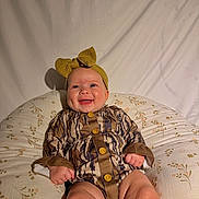 Averi joined the competition — help win amazing prizes! baby, child, infant, smiling, portrait, indoor, seated, bow, headband, outfit, patterned_clothing, buttons, floral_cushion, bed, fabric, shadow, face, happy, photography, closeup