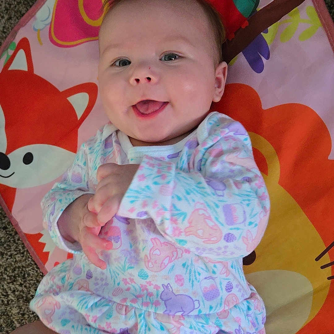 Taylin is registered to the contest to win money with this photo: animal_pattern, baby, carpet, child, clothing, colorful, cute, face, hands, happy, indoors, infant, lying_down, pastel_colors, play_mat, portrait, smiling, soft_light, tongue_out, young