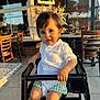 Milan is registered to the contest to win money with this photo: casual, chair, child, cute, expression, face, floor, furniture, green_shorts, high_chair, indoor, person, portrait, reflection, restaurant, sunlight, table, toddler, white_shirt, window