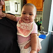 Leilani is registered to the contest to win money with this photo: baby, infant, person, holding, kitchen, cabinet, microwave, bib, pink_clothing, smile, face, drool, adult, glasses, window, countertop, food_container, arm, hand, indoor