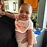 baby, infant, person, holding, kitchen, cabinet, microwave, bib, pink_clothing, smile, face, drool, adult, glasses, window, countertop, food_container, arm, hand, indoor