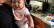 Leilani joined the competition — help win amazing prizes! baby, child, person, smiling, indoor, kitchen, bib, minnie_mouse, adult, holding, wooden_cabinet, microwave, stove, countertop, pot, phone, food_container, tile_floor, glass, happy