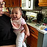 baby, child, person, smiling, indoor, kitchen, bib, minnie_mouse, adult, holding, wooden_cabinet, microwave, stove, countertop, pot, phone, food_container, tile_floor, glass, happy